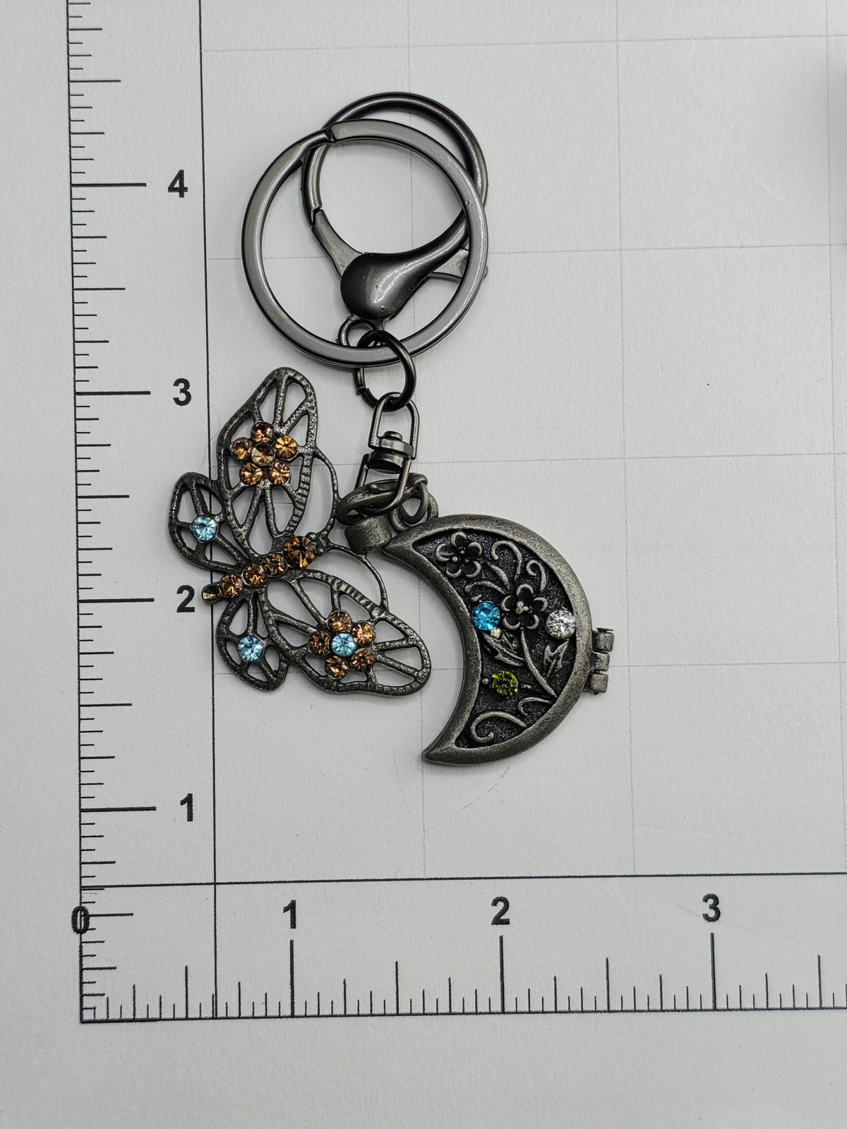 A decorative keychain with a butterfly and a moon charm, placed on a ruler for size comparison.