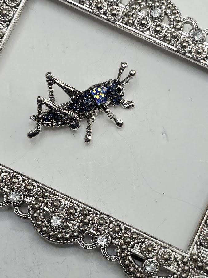 A silver brooch in the shape of a grasshopper, adorned with blue and yellow gemstones, is displayed on a white surface.