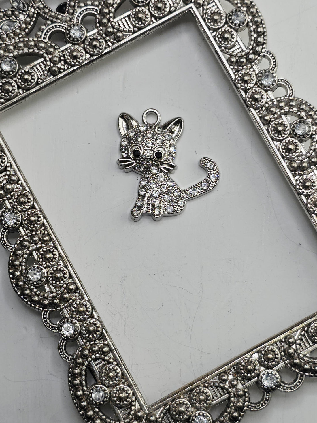 A silver-colored cat charm with sparkling rhinestones is displayed on a white surface, surrounded by an ornate, decorative metal frame.