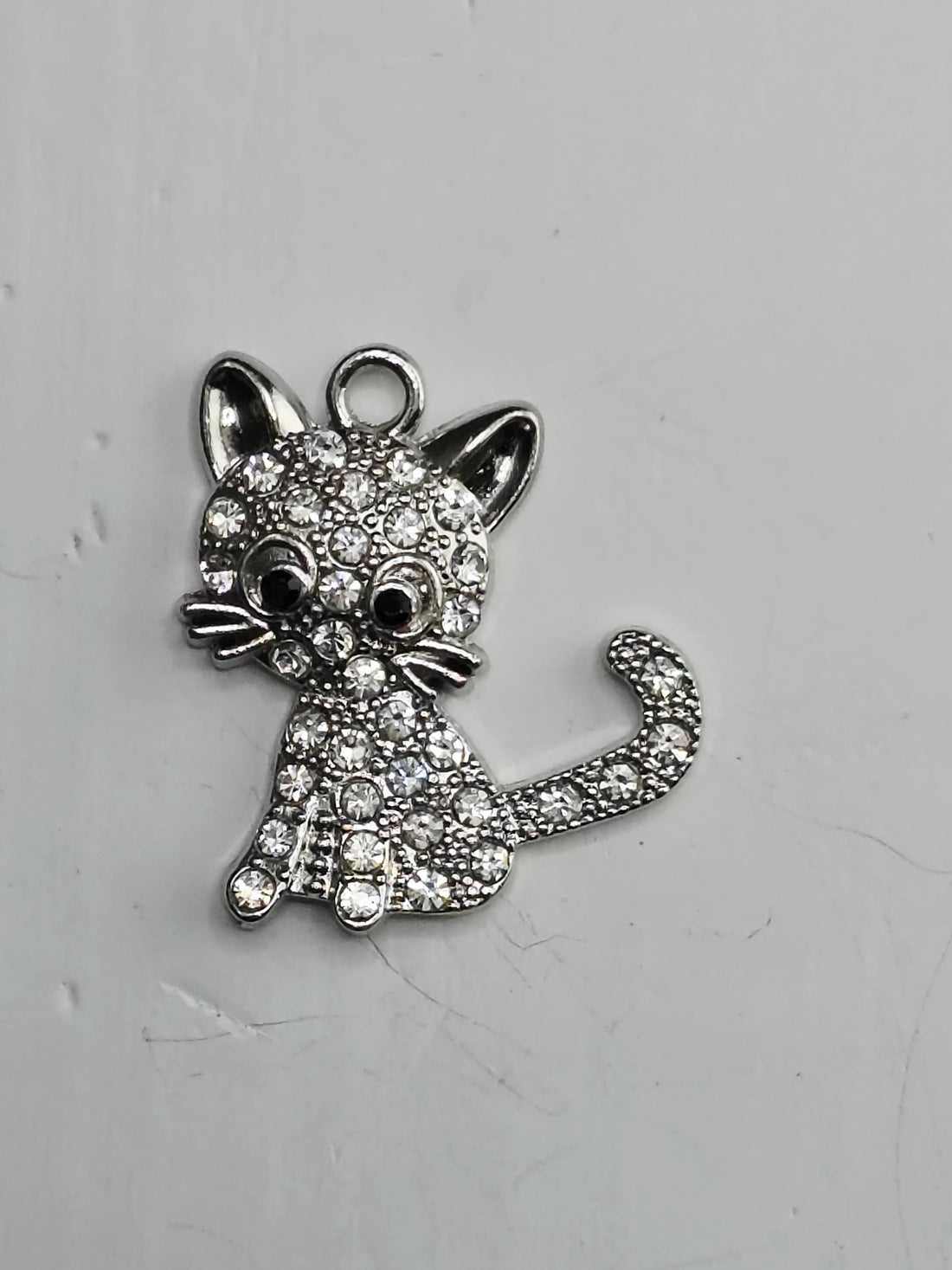 A silver-colored cat charm with sparkling rhinestones is displayed on a white surface, surrounded by an ornate, decorative metal frame.