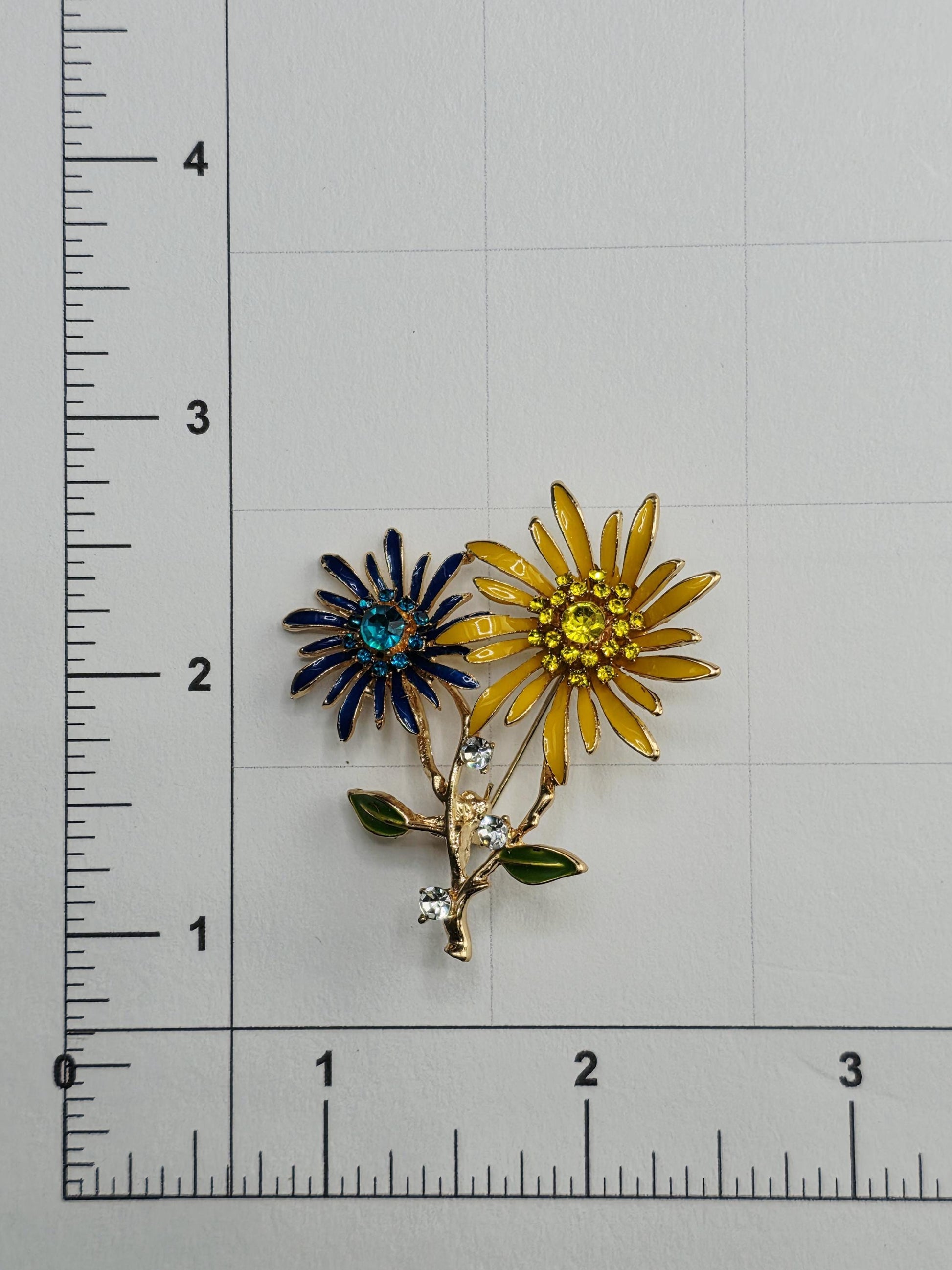 a decorative brooch with two yellow flowers and a blue flower, placed on a white surface next to a ruler for scale.
