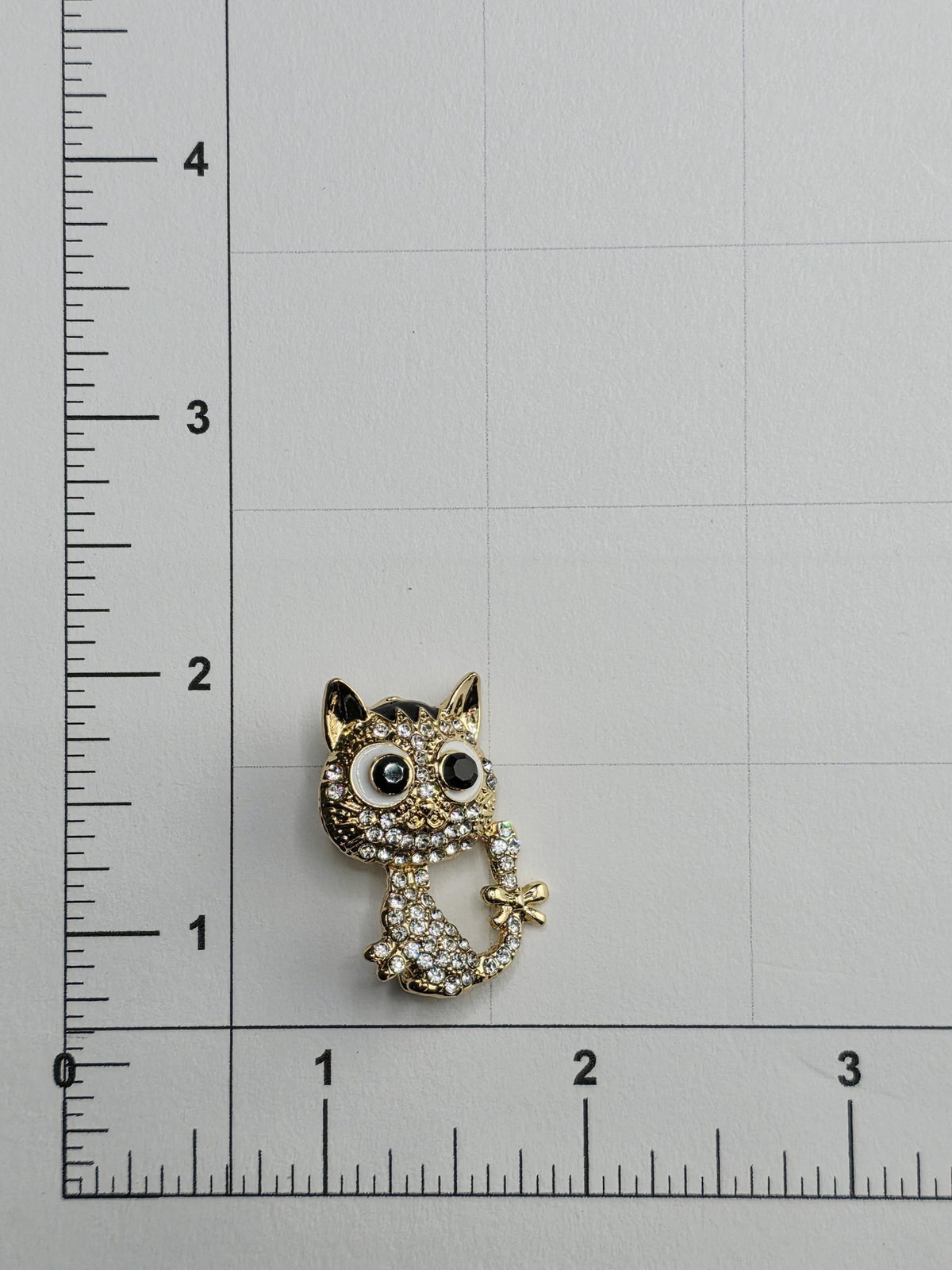 a small, ornate cat-shaped brooch with rhinestones or crystals on it, placed on a ruler for size comparison.