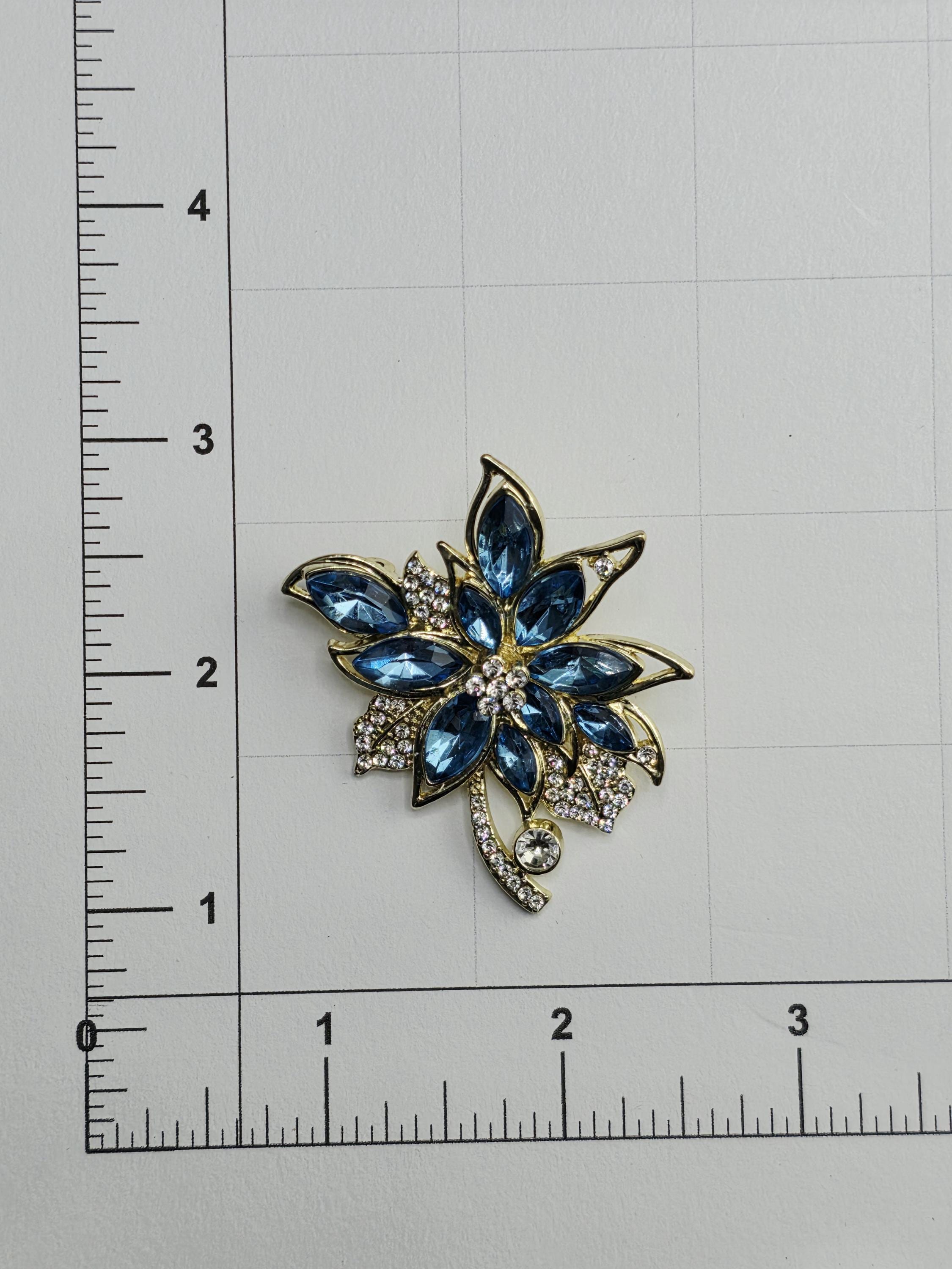 A decorative brooch with blue gemstones and a gold-colored design, placed on a ruler for size reference.