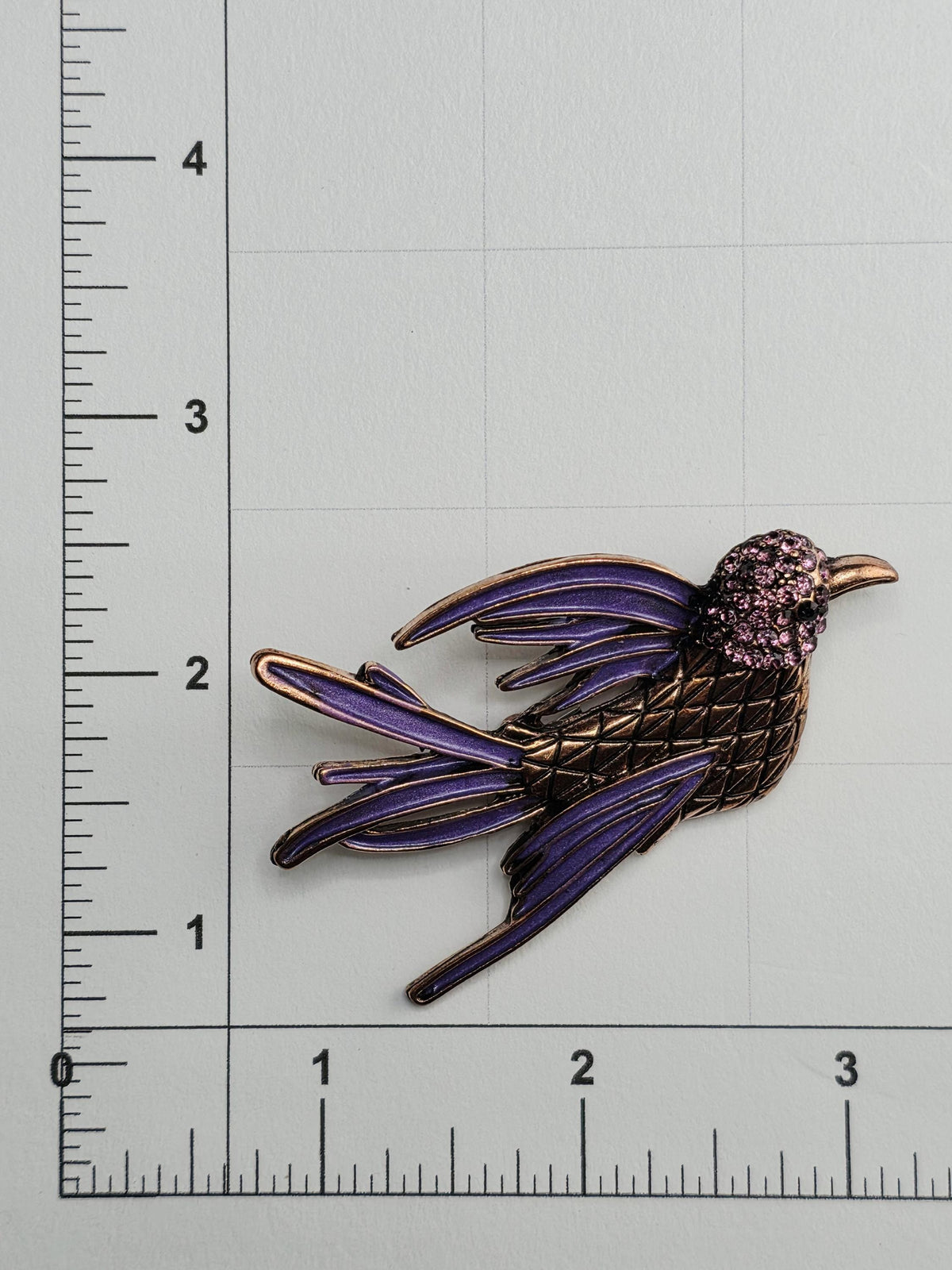 a decorative bird brooch with a purple and gold color scheme, placed on a ruler for size reference.