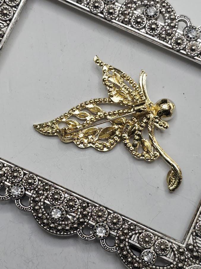 A decorative butterfly brooch with a pearl at the center, surrounded by sparkling rhinestones, is displayed on a white surface within an ornate frame.