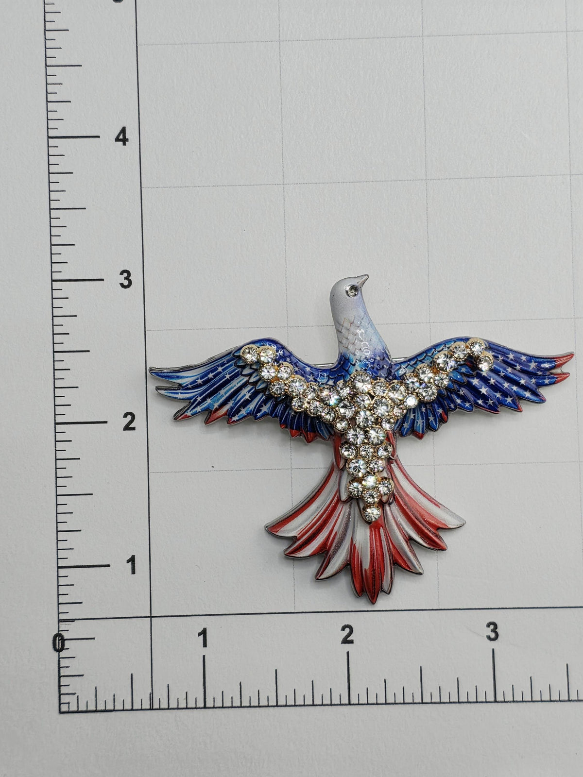 a decorative eagle brooch with a blue and red color scheme, adorned with rhinestones or crystals. The brooch is placed on a ruler for scale, indicating its size.