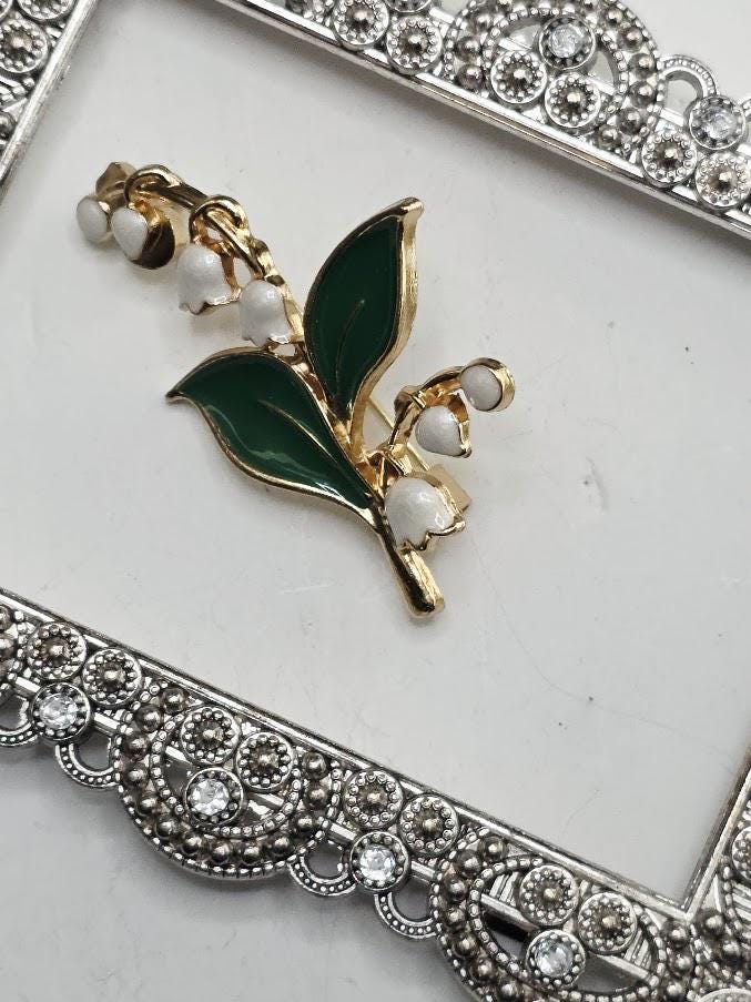 A decorative brooch with a green leaf and white flowers is displayed on a white surface, framed by a silver ornate frame.