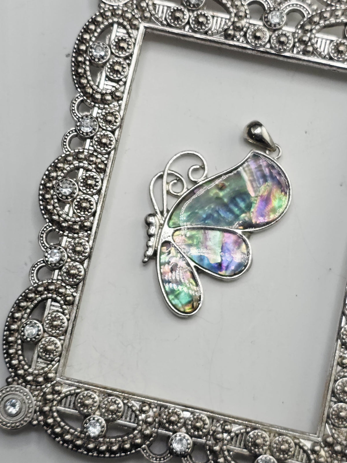 A silver butterfly pendant with a colorful iridescent pattern is displayed in a decorative silver frame.