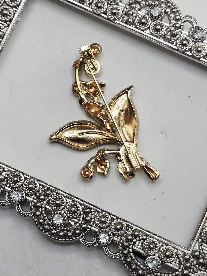 A gold brooch in the shape of a lily of the valley flower, with a leaf and flower design, is displayed on a white surface within a decorative metal frame.