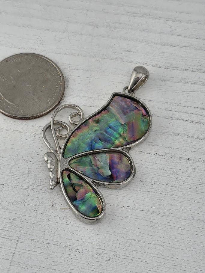 A silver-toned pendant in the shape of a butterfly, with colorful iridescent inlays, is shown next to a coin on a wooden surface.