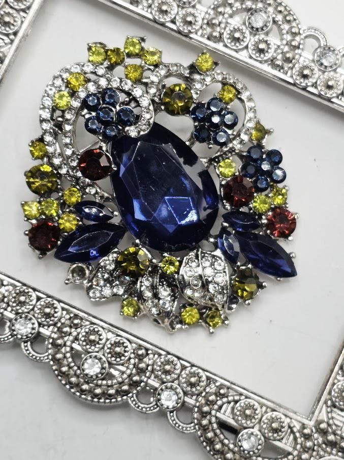 a close-up view of a decorative piece of jewelry, which appears to be a brooch or pendant. The piece is adorned with a variety of colorful gemstones, including blue, green, yellow, and red stones, as well as some metallic elements. The jewelry piece is set within an ornate, silver-colored frame, which adds to its overall aesthetic appeal.