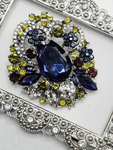 a close-up view of a decorative piece of jewelry, which appears to be a brooch or pendant. The piece is adorned with a variety of colorful gemstones, including blue, green, yellow, and red stones, as well as some metallic elements. The jewelry piece is set within an ornate, silver-colored frame, which adds to its overall aesthetic appeal.