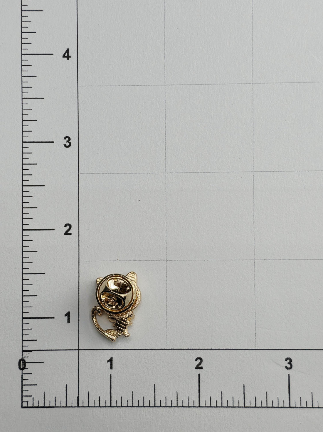 a small gold-colored pin or clip placed on a white surface, with a ruler for scale.