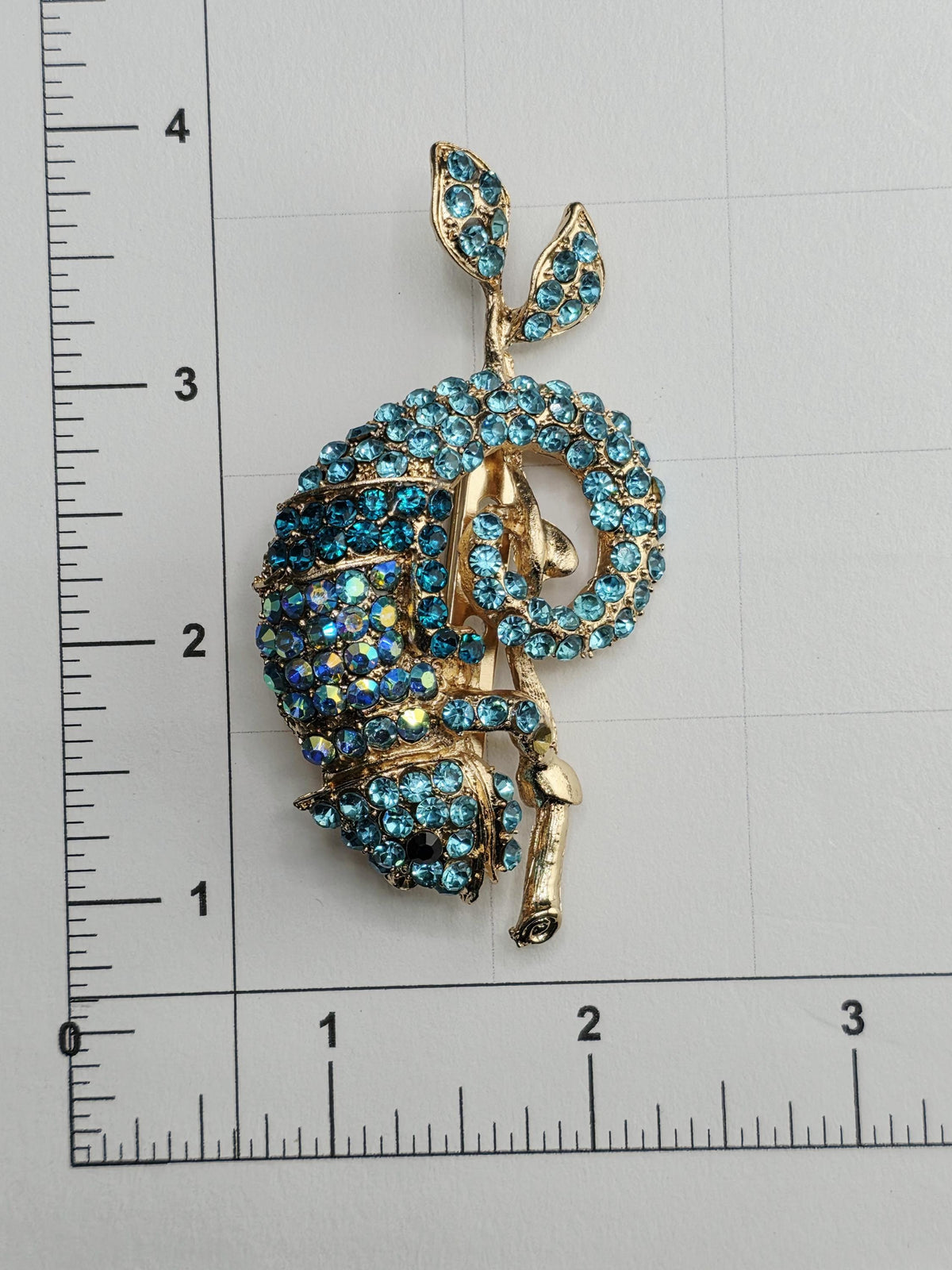 A decorative brooch with blue gemstones and a gold leaf design, placed on a ruler for size comparison.