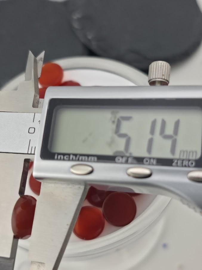 A digital caliper is measuring the diameter of red objects, which appear to be gemstones or beads. The caliper is displaying the measurement as 5.14 millimeters.