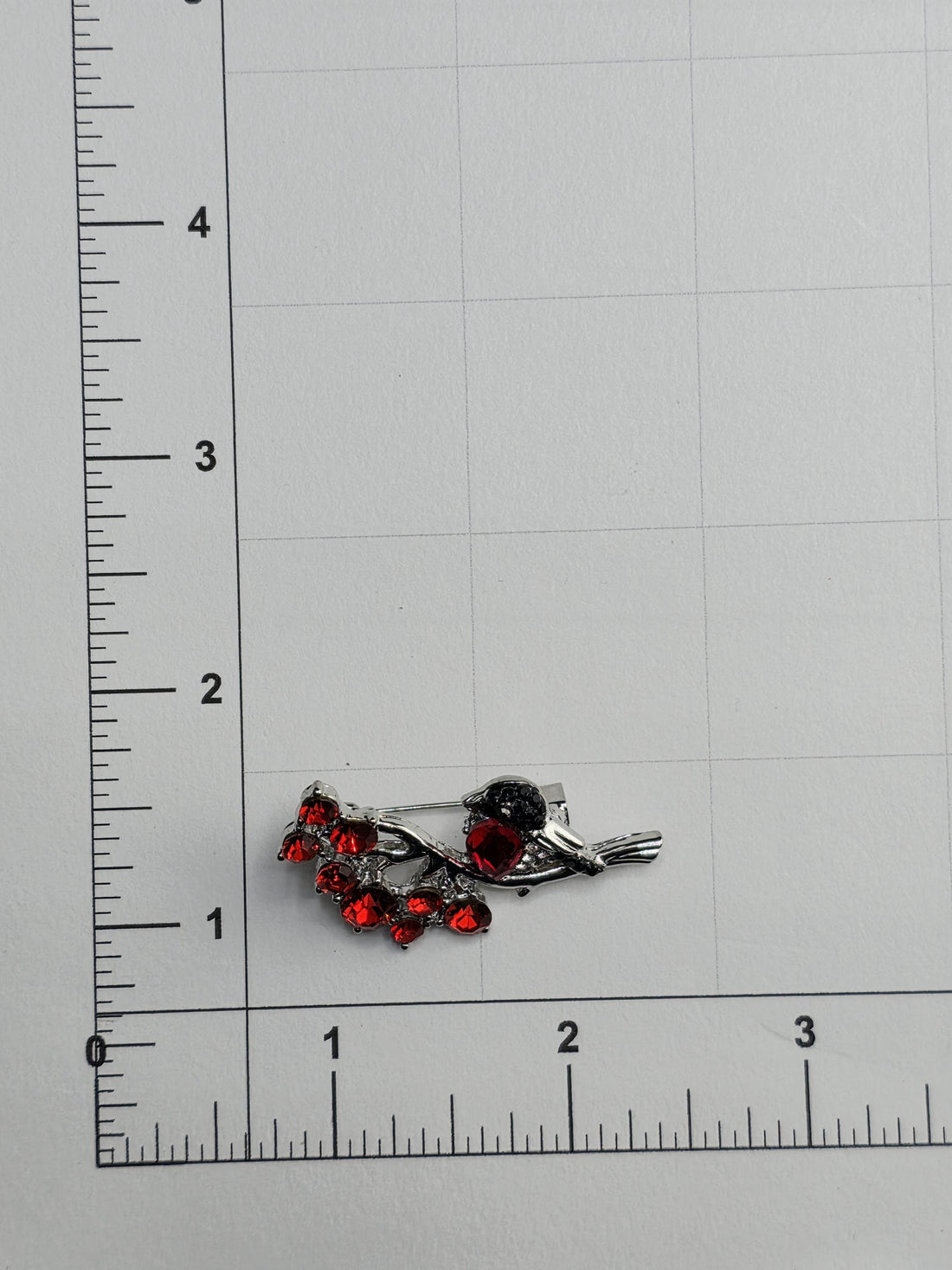 a silver brooch with red gemstones, placed on a white surface next to a ruler for size comparison.