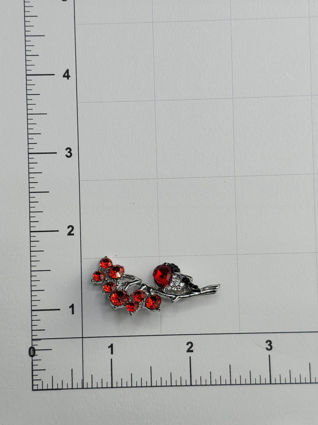 a silver brooch with red gemstones, placed on a white surface next to a ruler for size comparison.