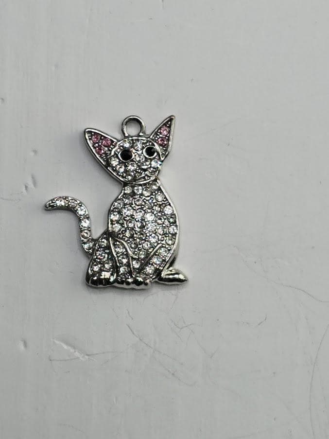 A silver-colored charm in the shape of a cat, adorned with small diamonds or crystals, is displayed against a plain white background.