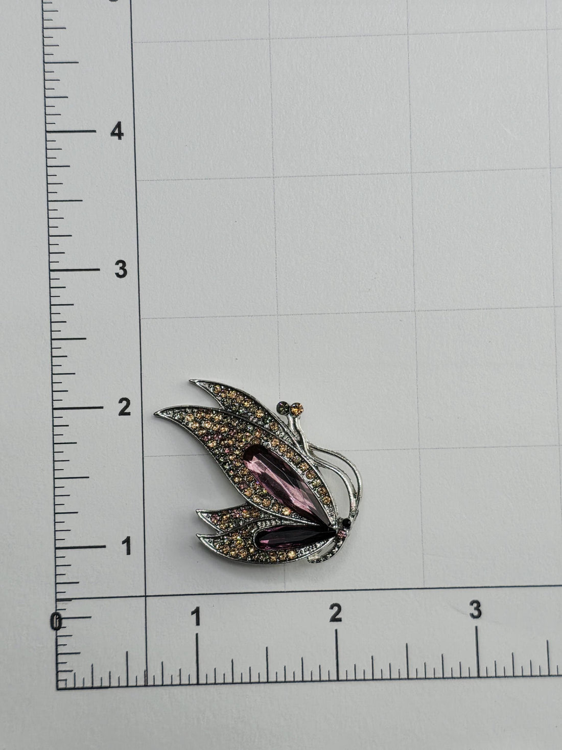 a decorative piece with a floral design and colorful gemstones, placed on a ruler for size comparison.