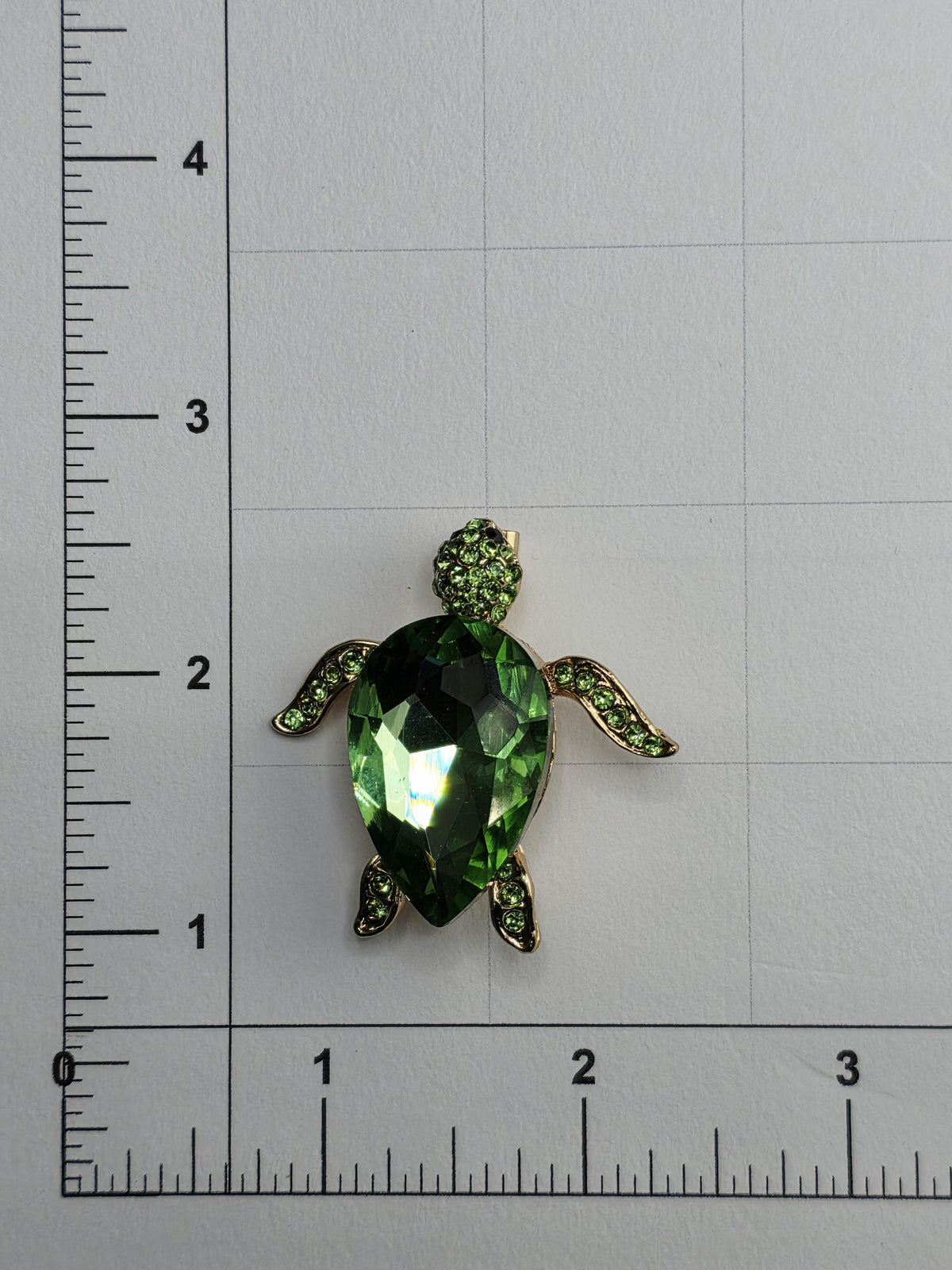 A green gemstone turtle brooch is placed on a ruler for size comparison.
