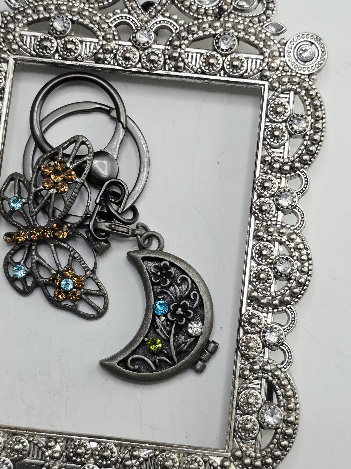 a silver-colored butterfly-shaped keychain with a moon-shaped charm attached to it, both adorned with colorful gemstones.