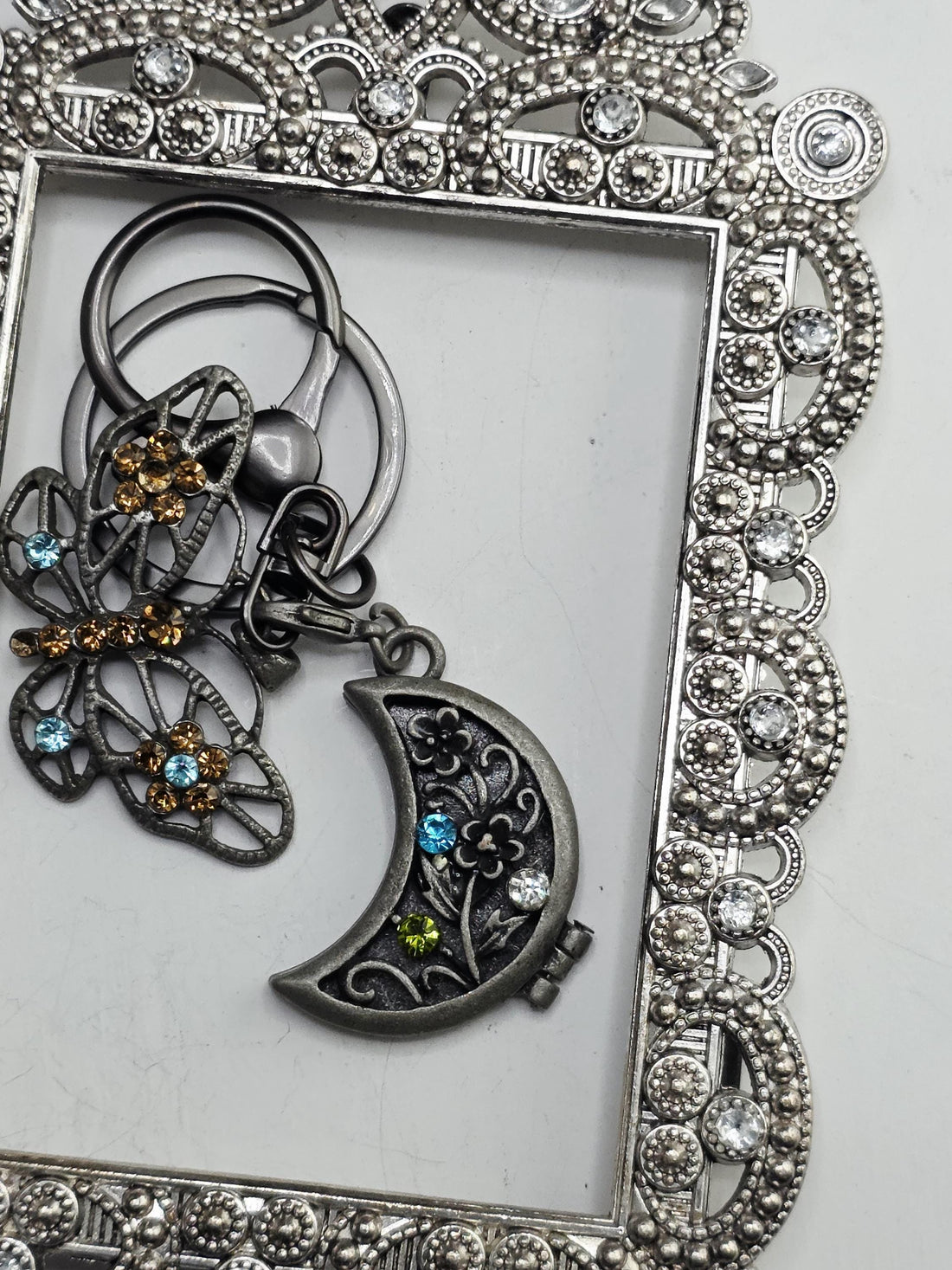 a silver-colored butterfly-shaped keychain with a moon-shaped charm attached to it, both adorned with colorful gemstones.