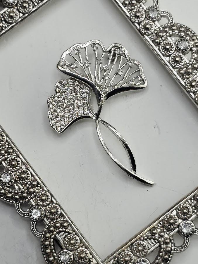 A silver-colored brooch with a leaf-like design and sparkling crystal elements is displayed against a white background.