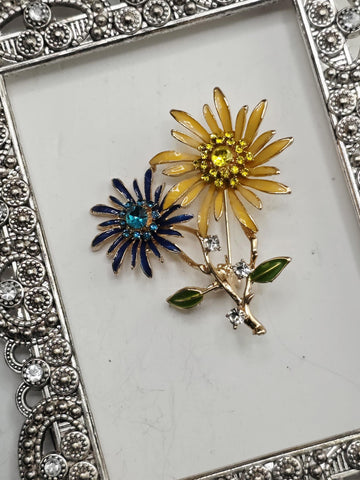 a decorative silver frame with a floral design, containing two colorful brooches with flowers and gemstones.