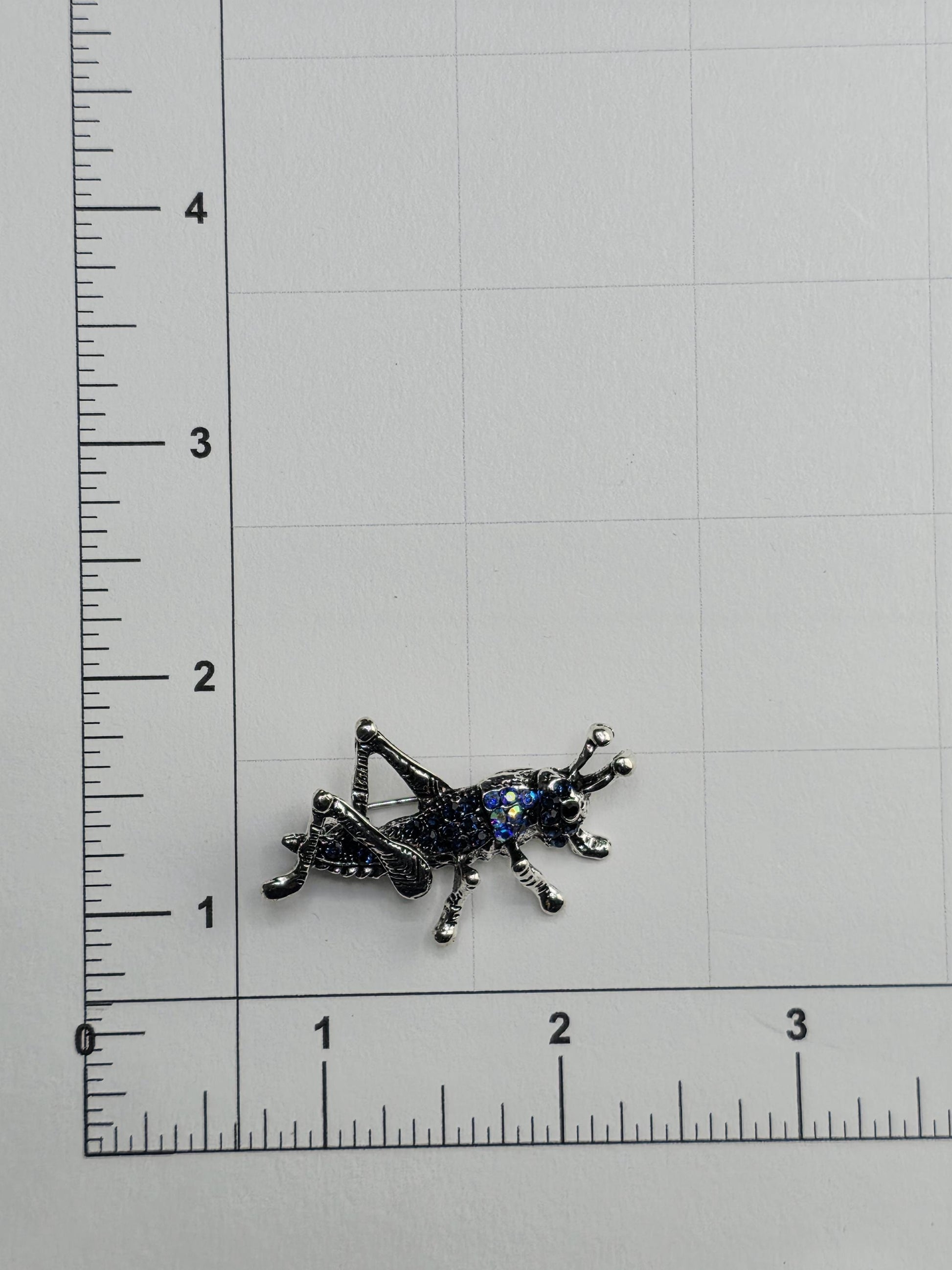 a shiny, metallic insect brooch placed on a white surface next to a ruler for size comparison.