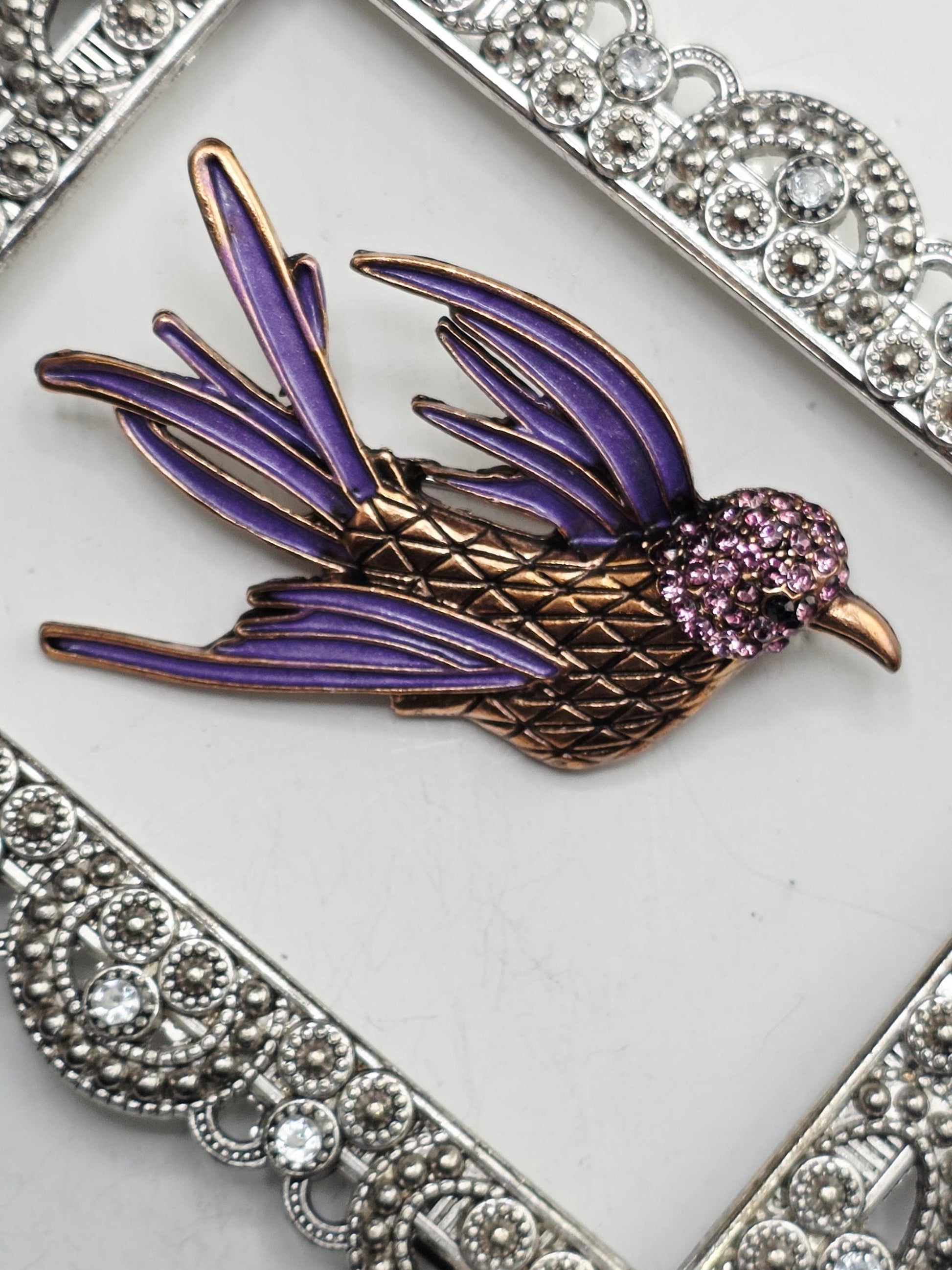 A decorative bird brooch with a purple body and gold accents, featuring a sparkling purple head and wings, is displayed on a white surface.