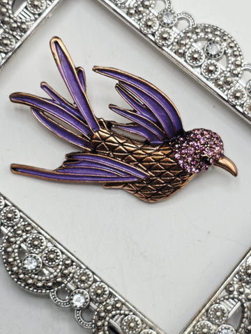 A decorative bird brooch with a purple body and gold accents, featuring a sparkling purple head and wings, is displayed on a white surface.