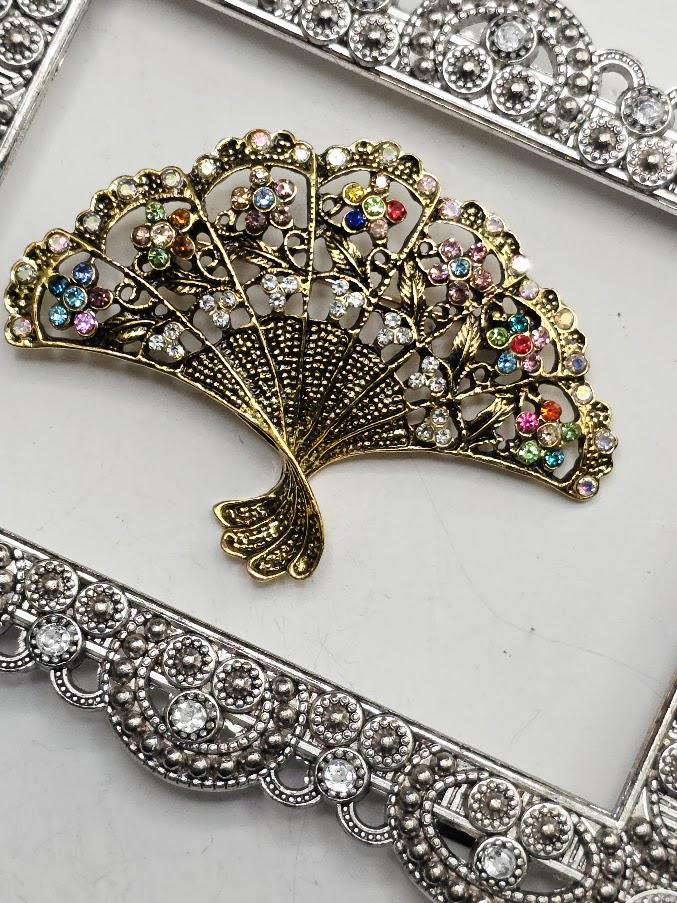 a decorative fan-shaped brooch with a gold-colored metal frame and a floral design, adorned with colorful gemstones.