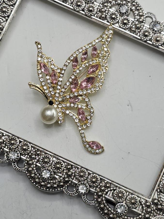 A decorative butterfly brooch with a pearl at the center, surrounded by sparkling rhinestones, is displayed on a white surface within an ornate frame.