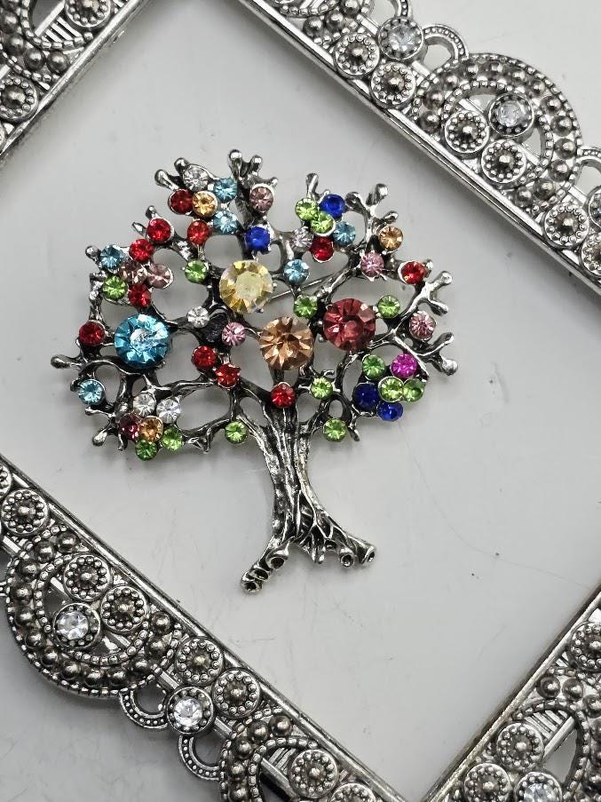 A decorative tree brooch with a variety of colorful gemstones, including red, blue, green, yellow, and pink, is displayed on a white surface.
