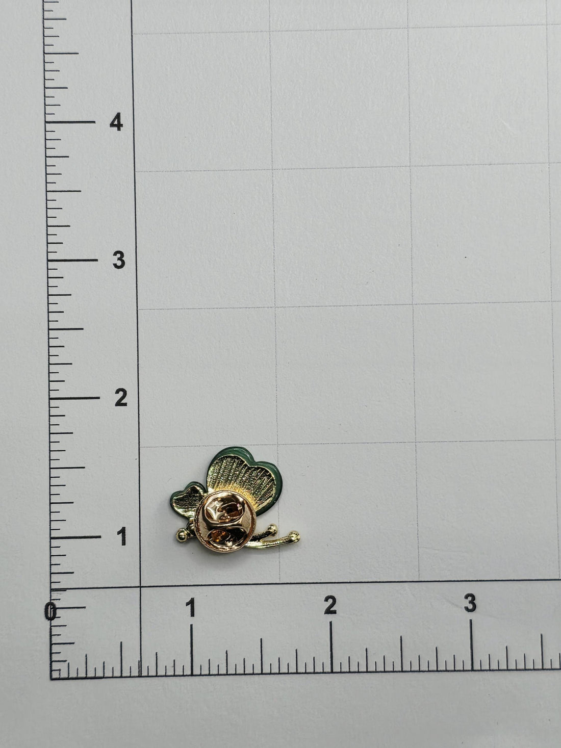 a small, gold-colored pin placed on a white surface next to a ruler for size comparison.