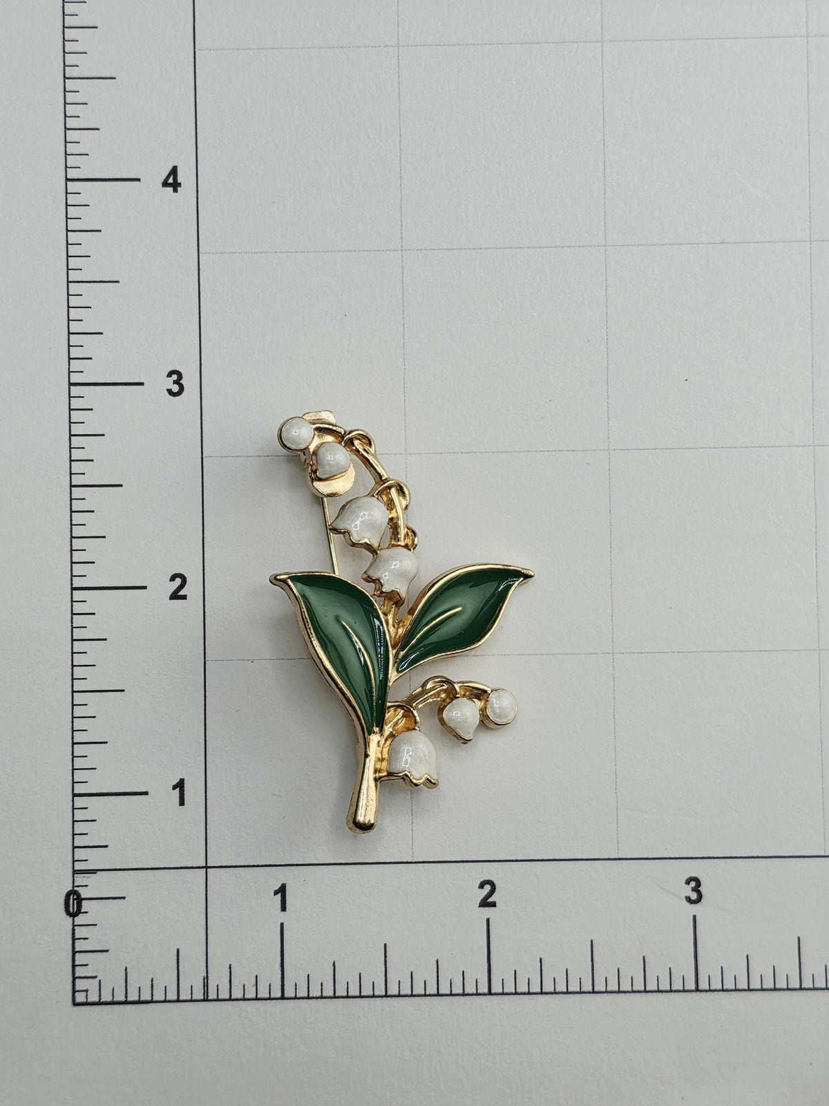 A decorative brooch in the shape of a lily of the valley flower, with green leaves and white flowers, is placed on a ruler for size comparison.