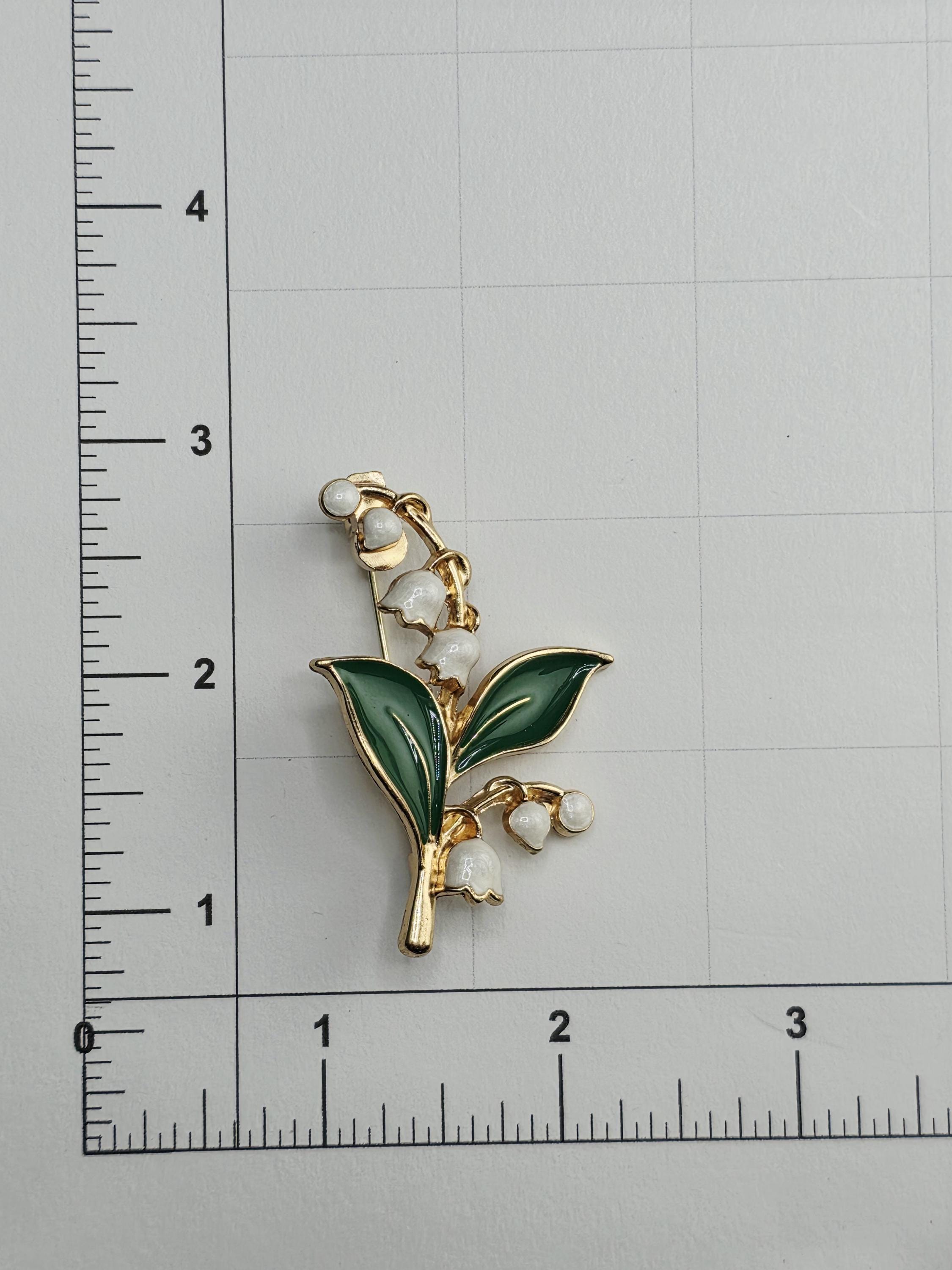 A decorative brooch in the shape of a lily of the valley flower, with green leaves and white flowers, is placed on a ruler for size comparison.