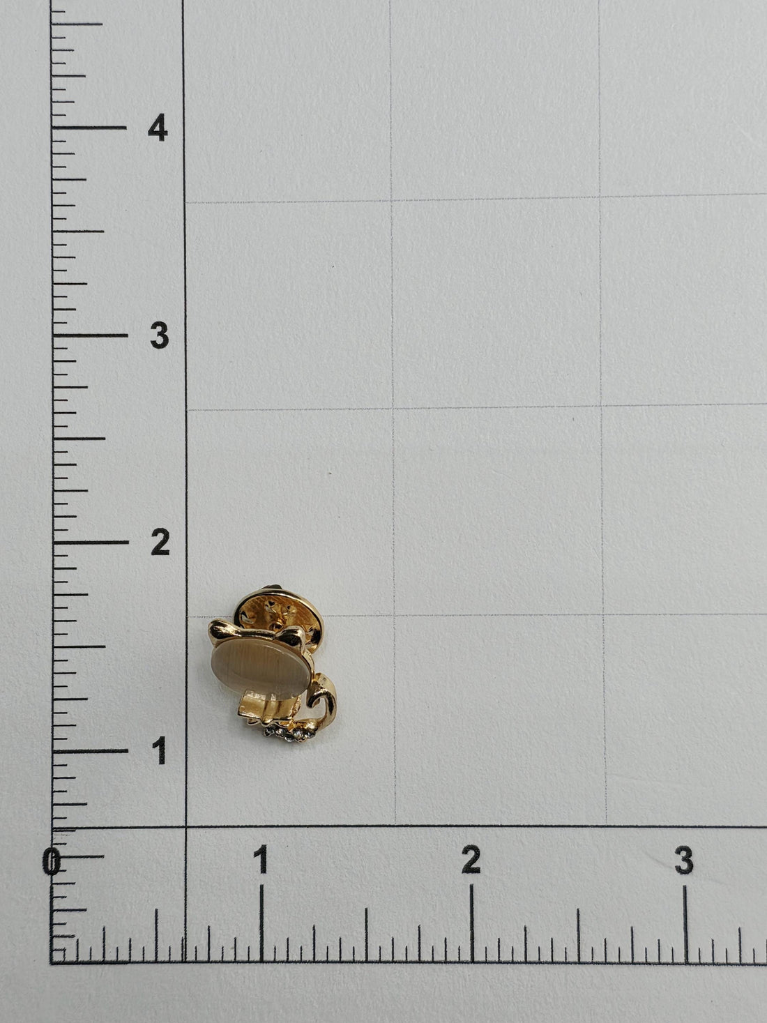 a small gold-colored pin or clip placed on a white surface, with a ruler for scale.