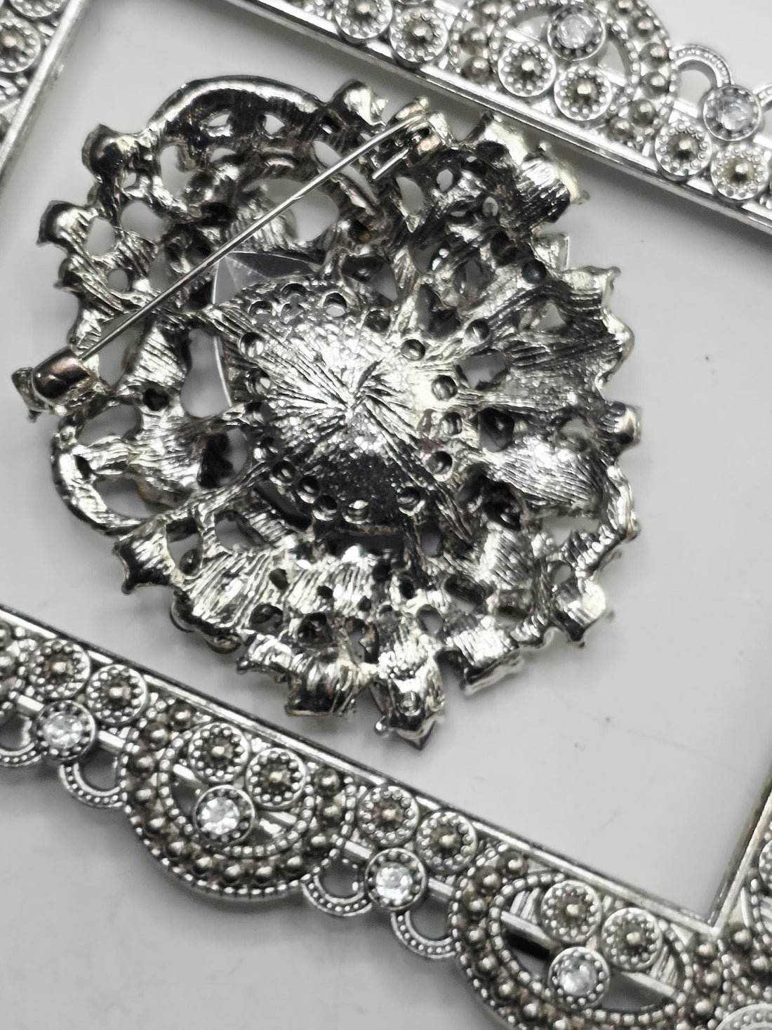 a close-up view of a decorative piece of jewelry, which appears to be a brooch or pendant. The piece is adorned with a variety of colorful gemstones, including blue, green, yellow, and red stones, as well as some metallic elements. The jewelry piece is set within an ornate, silver-colored frame, which adds to its overall aesthetic appeal.
